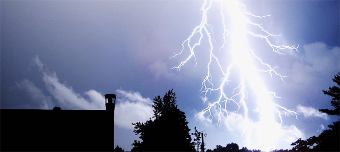 Photo of lightning bolt in the night sky over trees and rooftops.